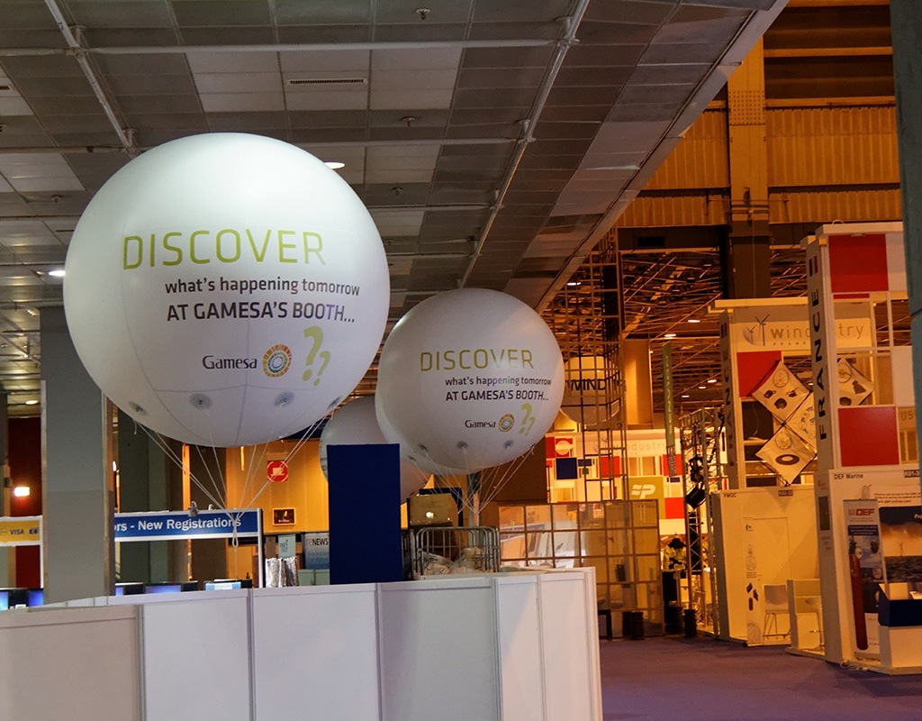 Ballons hélium publicitaires visibles au-dessus de plusieurs stands lors d’un salon.